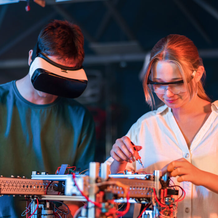 Teens doing experiments in robotics in a laboratory. Boy in VR headset and girl in protective glasses working with a robot. Red and blue illumination
